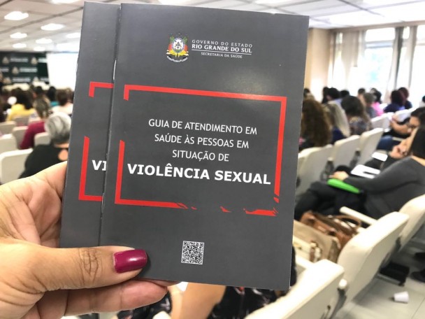 Uma mão feminina segura o Guia de Atendimento, que tem a capa preta.